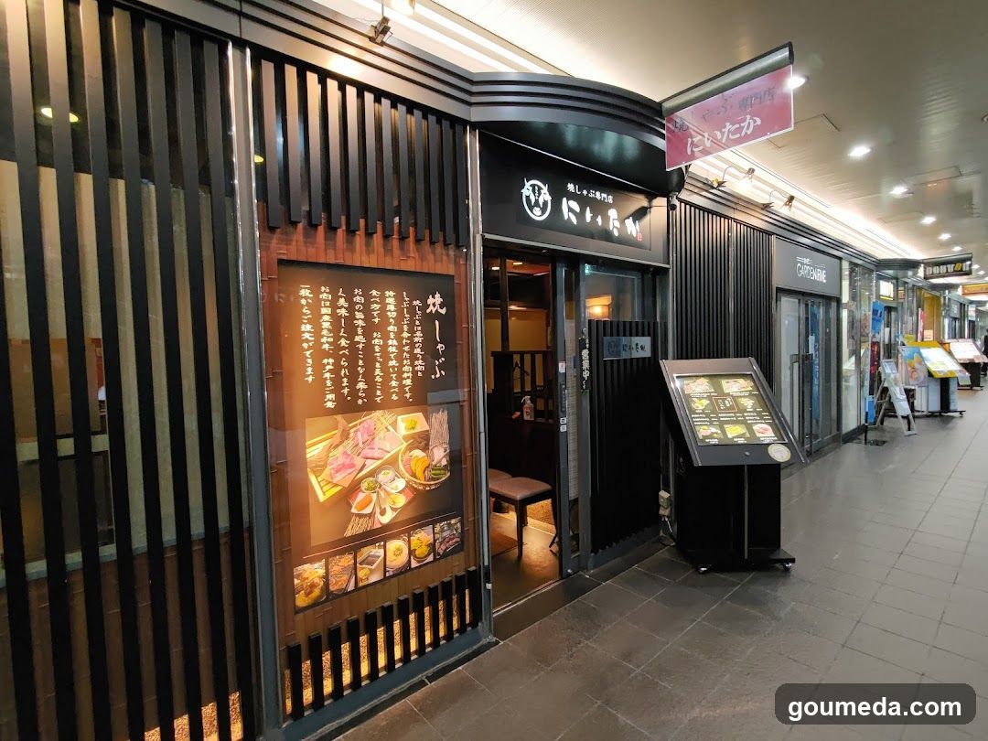 The image shows the exterior of a restaurant, featuring a black facade with vertical slats and illuminated signage in Japanese characters. A large poster on the window displays food items, including grilled meat and sides. To the right, there is a digital menu board stand showing various dishes with prices. The entrance has glass doors leading into the interior.
