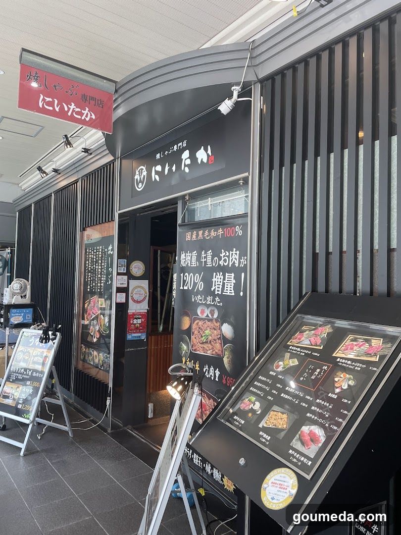 The image shows the exterior of a Japanese yakitori shop called 'Niitaka' (にいたか). The storefront features black vertical slats and signage in both English and Japanese, including a red banner reading 'Yakiyabu Niitaka'. A large menu board is displayed outside with images of grilled skewers and prices listed in yen. There are additional signs advertising beef products and promotions inside the entrance area.
