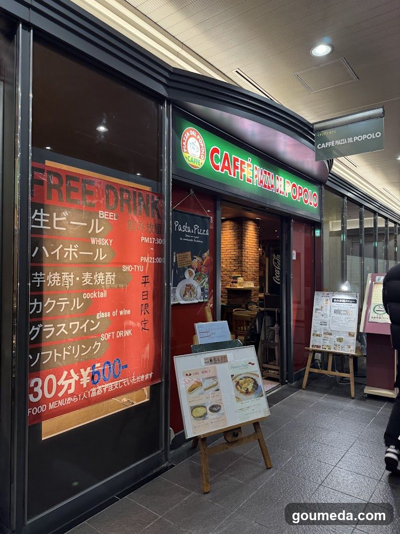 A photo of the physical exterior entrance and storefront facade of Caffè Pizza del Popolo, featuring visible signage on the building including a green illuminated sign with white lettering and a hanging directional sign. A large orange promotional poster advertises free drinks (beer, whiskey, cocktail, sake, wine, soft drink) from 30 minutes before closing at ¥600. Outside menu boards display food items like pasta pizza and other dishes with prices.