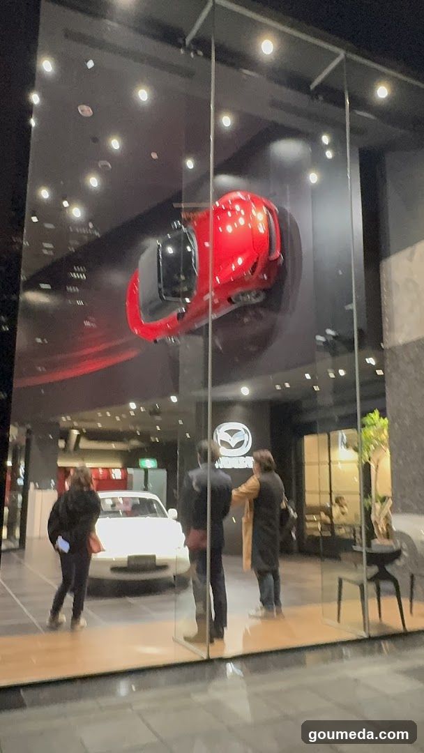 Night view of a Mazda dealership entrance featuring a large glass facade with an illuminated red sports car suspended above the entrance. Below, three people stand near a white sedan inside the showroom. The Mazda logo is visible on the wall behind them.