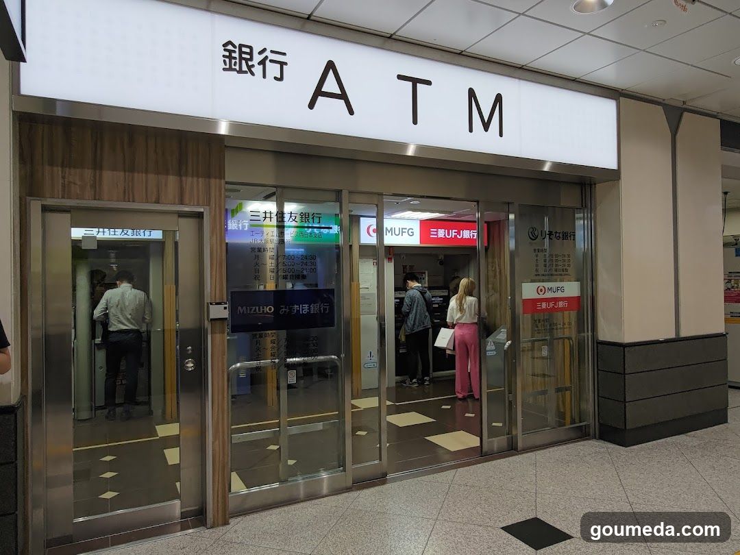 A modern bank ATM entrance with glass doors and signage displaying '銀行 ATM' (Bank ATM). Inside, customers are using ATMs from Mizuho and MUFG banks. The interior is well-lit with a clean, professional design.