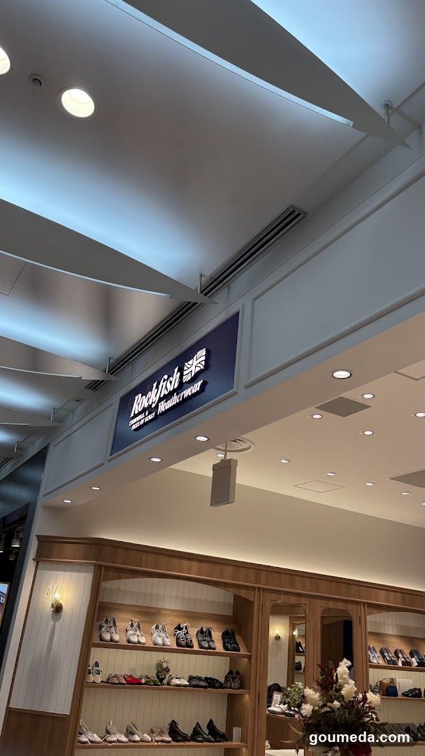 Interior view of a shoe store with wooden display shelves filled with various footwear including sneakers, boots, and heels. A blue sign above reads 'Rockfish Footwear' with a UK flag symbol. Lighting is bright with recessed ceiling lights.
