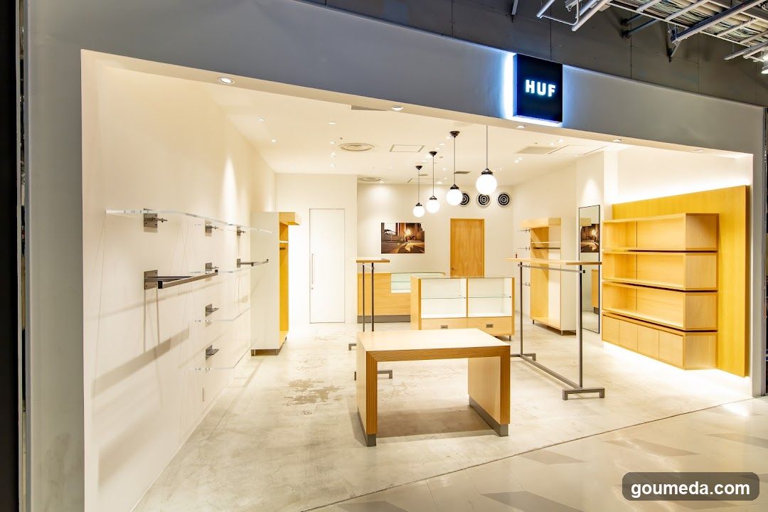 Interior view of a modern retail store with minimalist design, featuring light wood shelving units, glass display racks, and pendant lighting. The entrance is visible with the brand name 'HUF' illuminated in blue on the facade above the doorway.