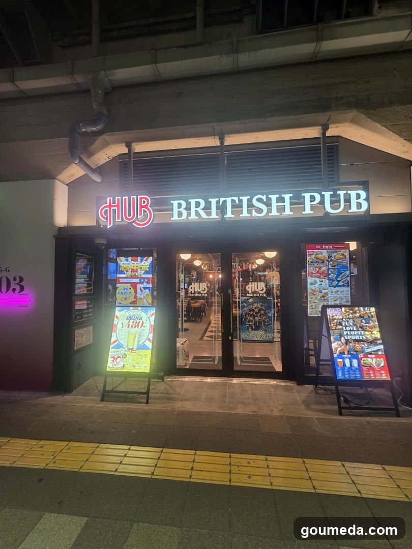 Night view of the storefront featuring illuminated signage reading 'HUB BRITISH PUB'. The entrance has glass doors with posters inside and outside, flanked by A-frame menu boards displaying food items and prices. Neon lights accentuate the sign above.