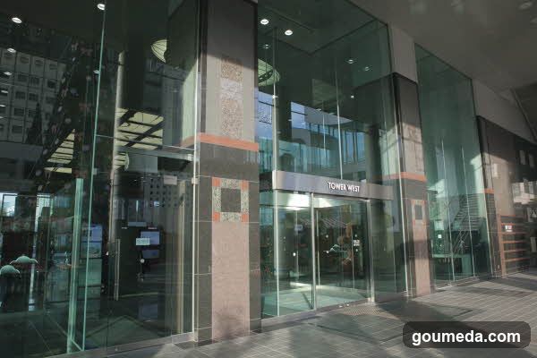 Modern glass facade with 'TOWER WEST' signage above entrance, reflecting urban surroundings.