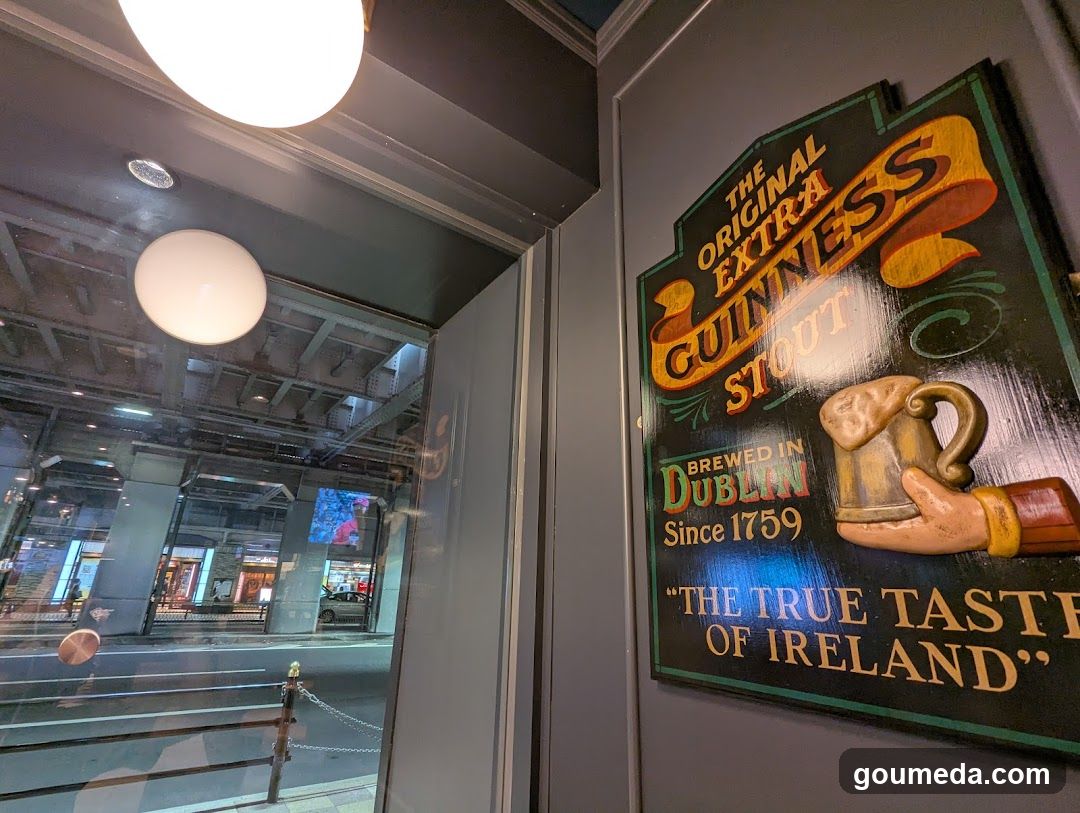 A close-up view of a shop entrance featuring a prominent Guinness Stout sign mounted on the wall. The sign reads 'The Original Extra Guinness Stout', 'Brewed in Dublin Since 1759', and 'The True Taste of Ireland'. Through the glass door, reflections of streetlights and urban surroundings are visible.