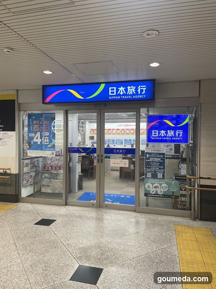 The image shows the entrance of a Nippon Travel Agency store, featuring glass doors and windows with signage displaying '日本旅行' (Japan Travel) in both Japanese and English. Inside, various promotional posters and brochures are visible on display racks.