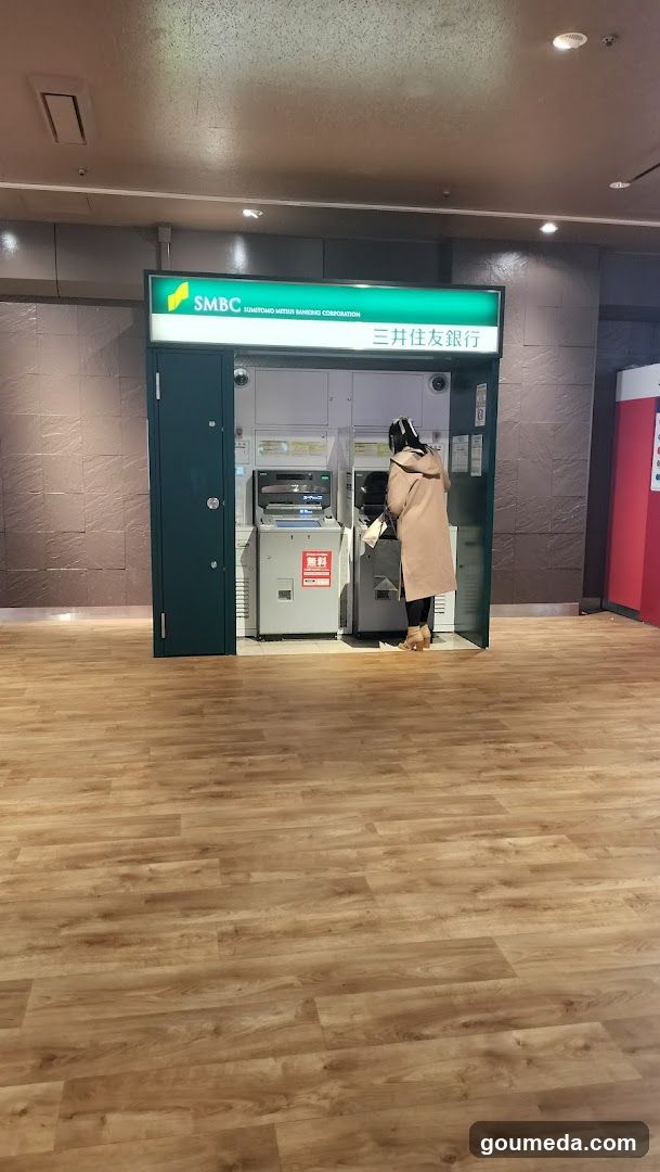 A self-service ATM booth with SMBC (Sumitomo Mitsui Banking Corporation) signage above the entrance. A person in a beige coat is standing at the machine, using it. The area has wooden flooring and tiled walls.