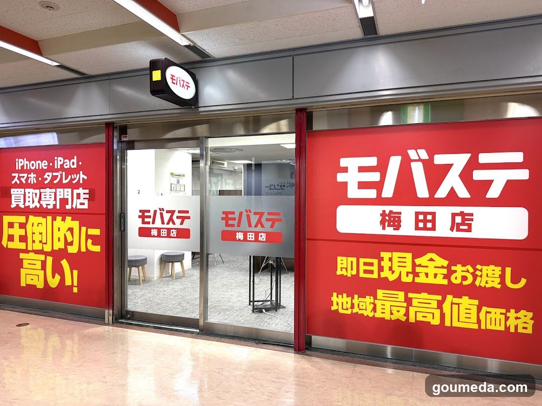 A storefront of Mobasite (モバステ) in the Umeda area, featuring large red signage with Japanese text advertising iPhone/iPad trade-in services and high cash payouts. The entrance has glass doors with the store name displayed on them.