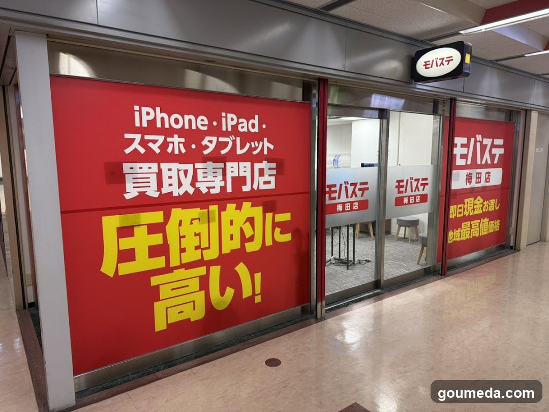 A shop front featuring a large red sign with white and yellow Japanese text advertising iPhone and iPad trade-ins, emphasizing high prices. The store is named 'Mobasite' (モバステ) and located in Umeda branch. Glass doors show the interior with chairs and tables.