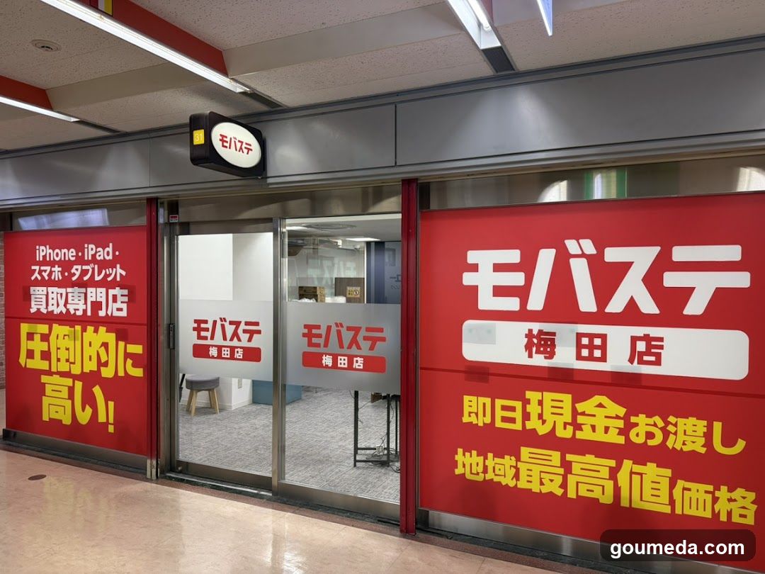 The image shows the storefront of a Mobasite (mobile phone trade-in) store located in the Meida area. The entrance features glass doors with 'Mobasite' and 'Meida Store' signage on them. Large red banners flank the entrance, advertising services like iPhone/iPad trade-ins, emphasizing high payout rates and immediate cash transfer. A hanging sign above reads 'Mobasite'. The interior is partially visible through the glass, showing office furniture.