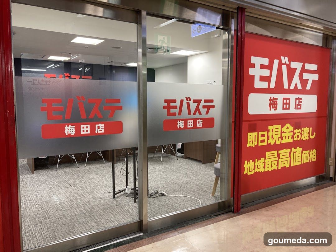 Glass storefront with red and white signage displaying 'モバステ' (Mobasite) and '梅田店' (Umeda Branch). Interior shows office furniture, carpeted floor, and equipment cables visible through the glass doors.